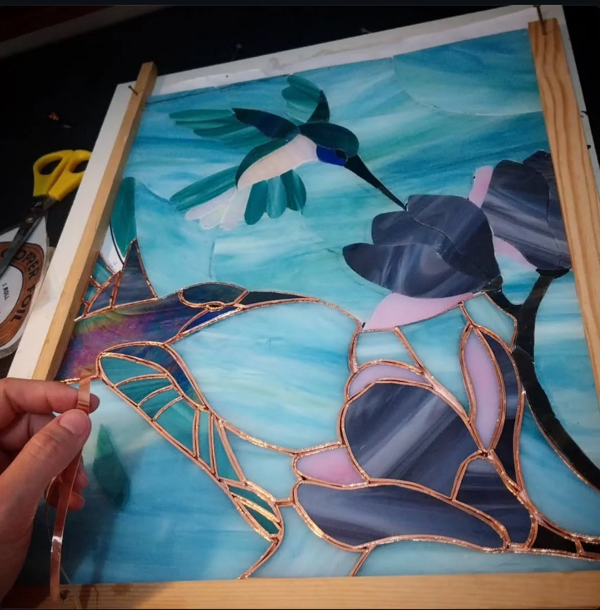 Vitral Tiffany de dos colibríes y magnolias en proceso, aplicando a mano la cinta de cobre a cada pieza de vidrio