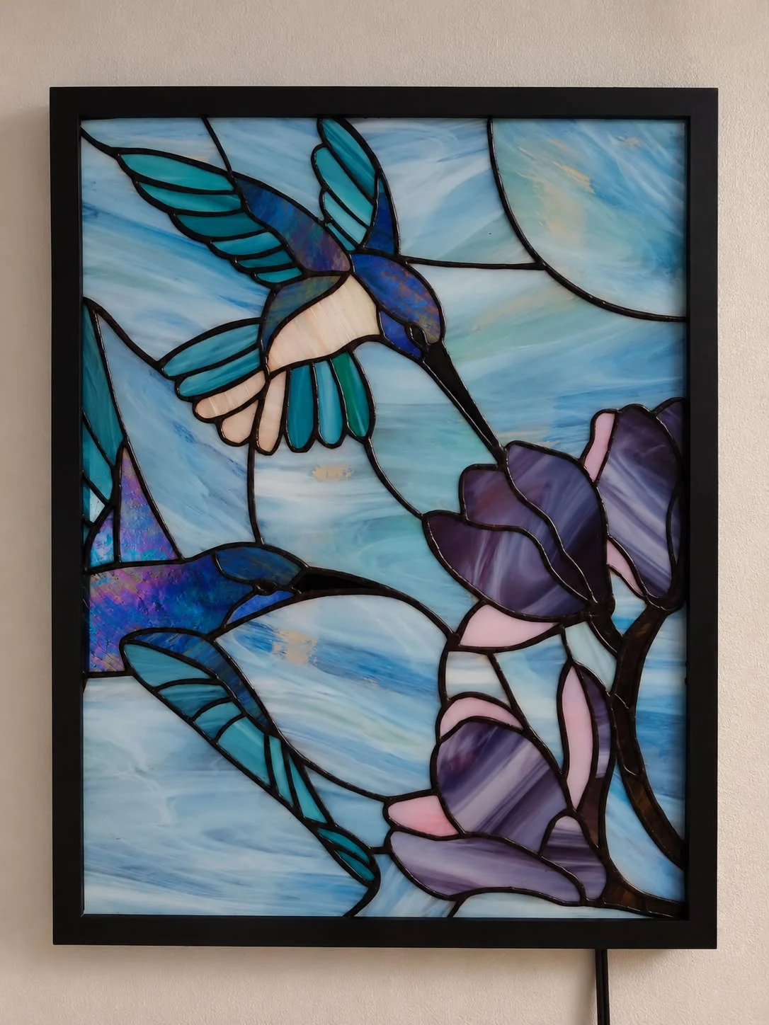 Vitral Tiffany de colibríes y magnolias terminado, en una caja de luz con marco negro colgada en la pared