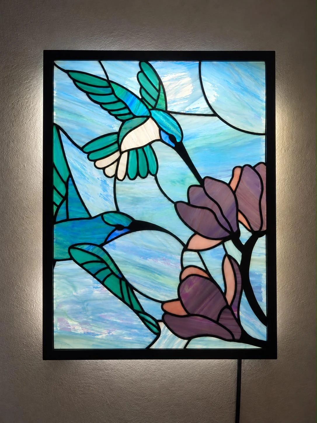 Vitral Tiffany de colibríes y magnolias retroiluminado en su caja de luz, con un halo de luz cálida sobre la pared